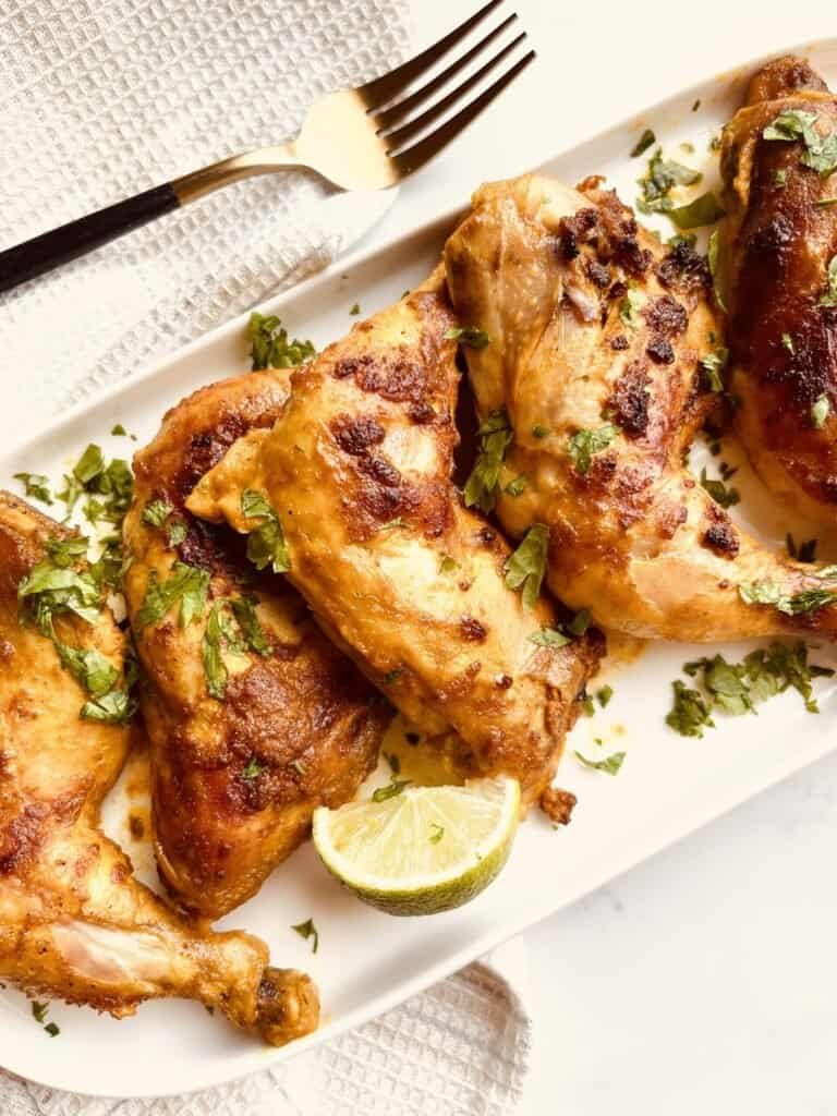 Grilled yogurt-marinated chicken pieces garnished with herbs and lime wedge on a white platter.