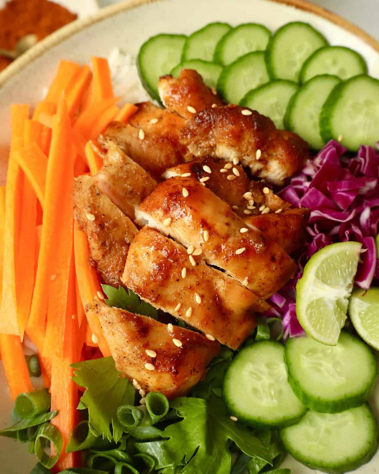 Grilled chili honey chicken with fresh cucumber, carrot, purple cabbage, and lime slices on a bowl.