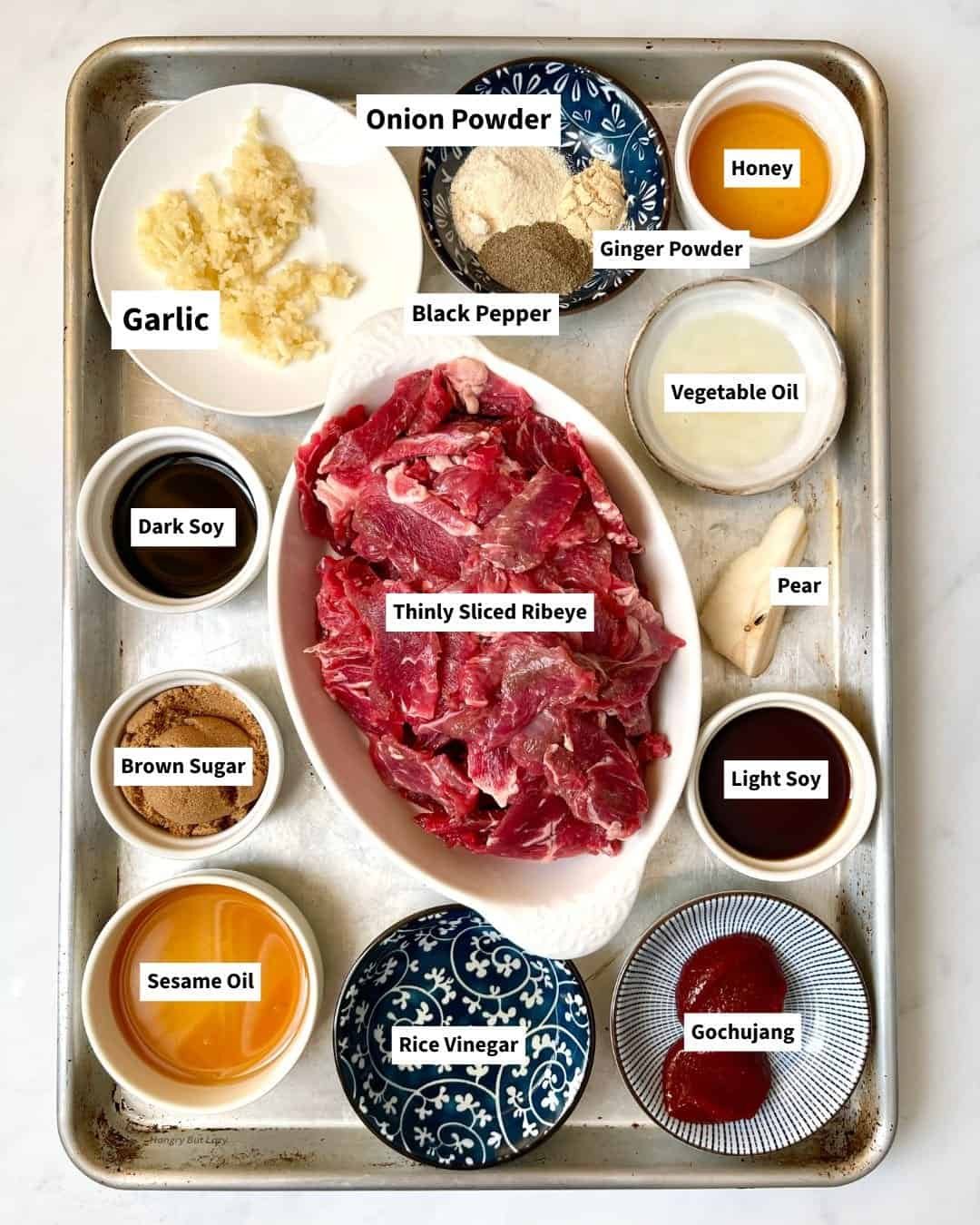 Gochujang beef marinade ingredients with thinly sliced ribeye and spices on a tray.