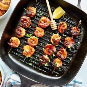 Grilled honey garlic shrimp skewers in a black grill pan with lemon wedge and fresh herbs.
