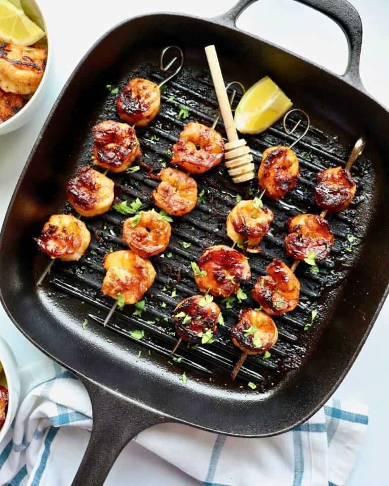 Grilled honey garlic shrimp skewers in a black grill pan with lemon wedge and fresh herbs.