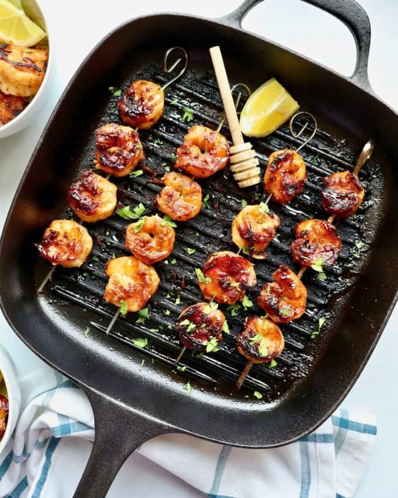 Grilled honey garlic shrimp skewers in a black grill pan with lemon wedge and fresh herbs.