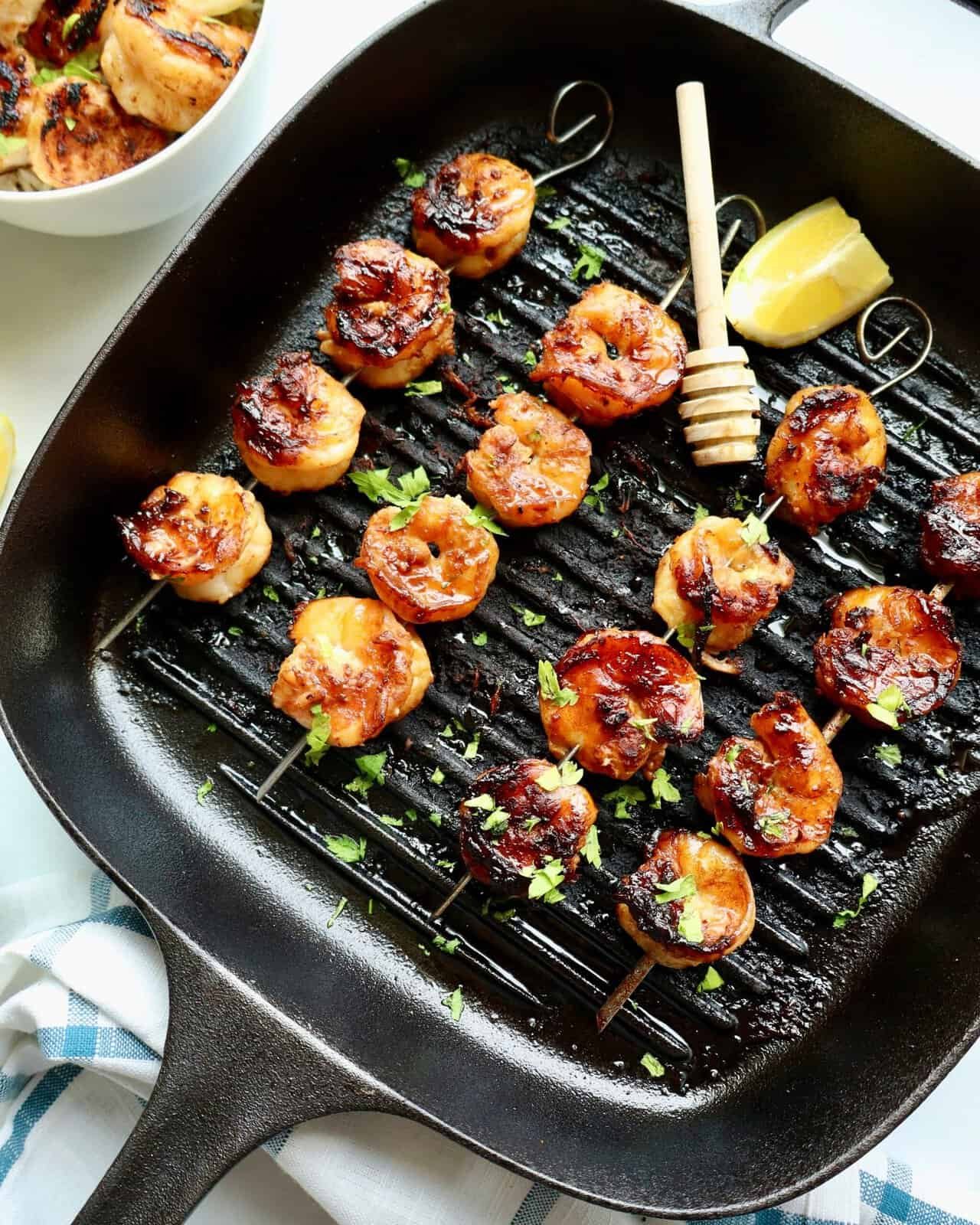 Grilled honey garlic shrimp skewers on a black grill pan with lemon wedge and honey dipper.
