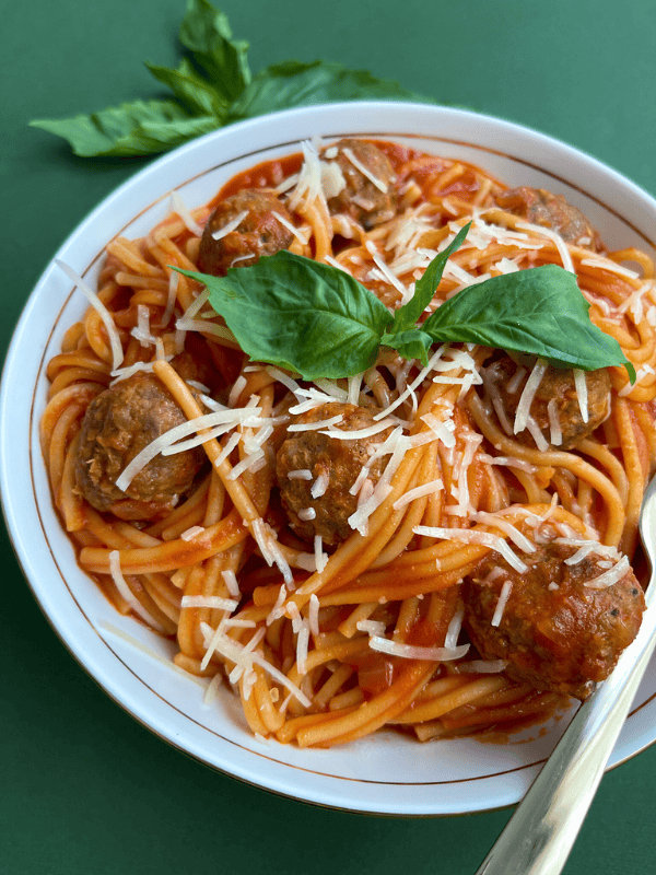 Spaghetti with meatballs, fresh basil, and grated cheese in a white bowl on a green background.