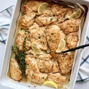 Juicy baked chicken thighs with lemon slices and fresh herbs in a white baking dish.