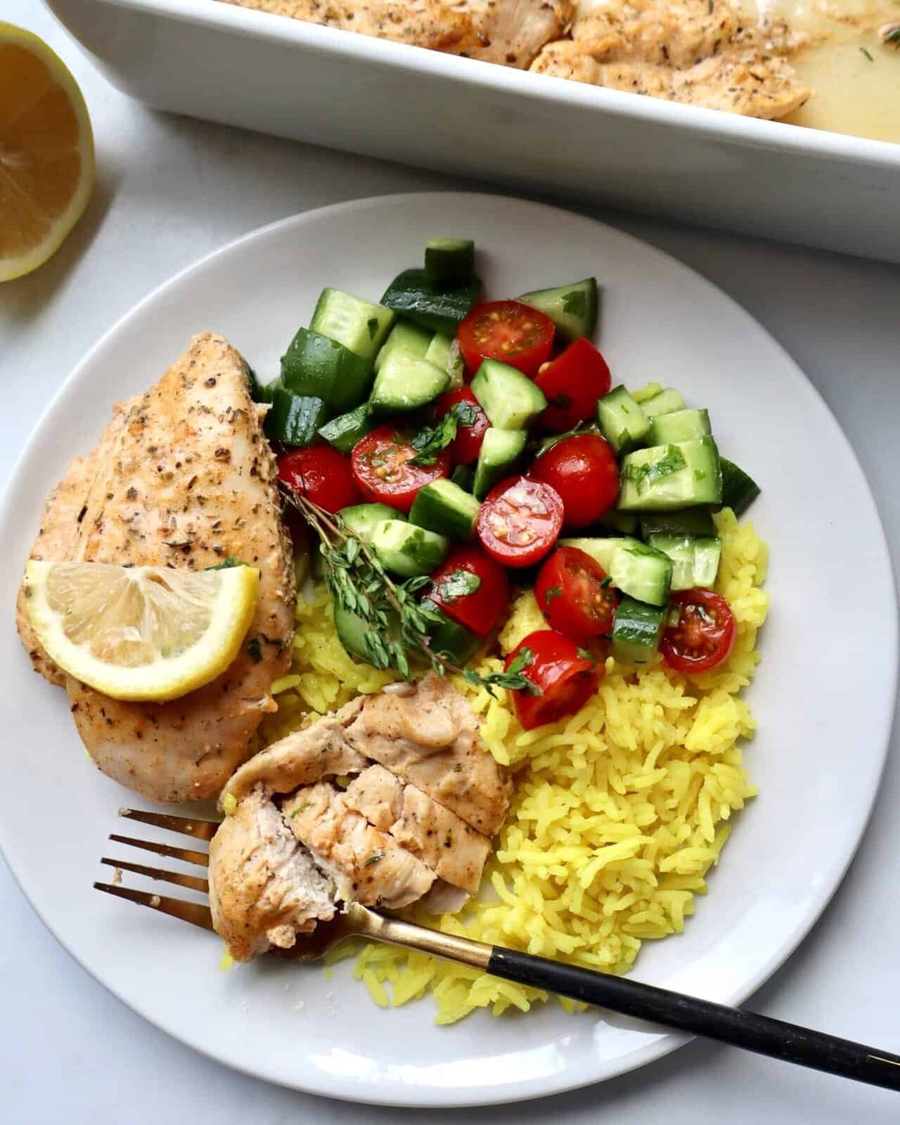 Grilled chicken with lemon, cucumber, cherry tomato salad, and yellow rice on a white plate.