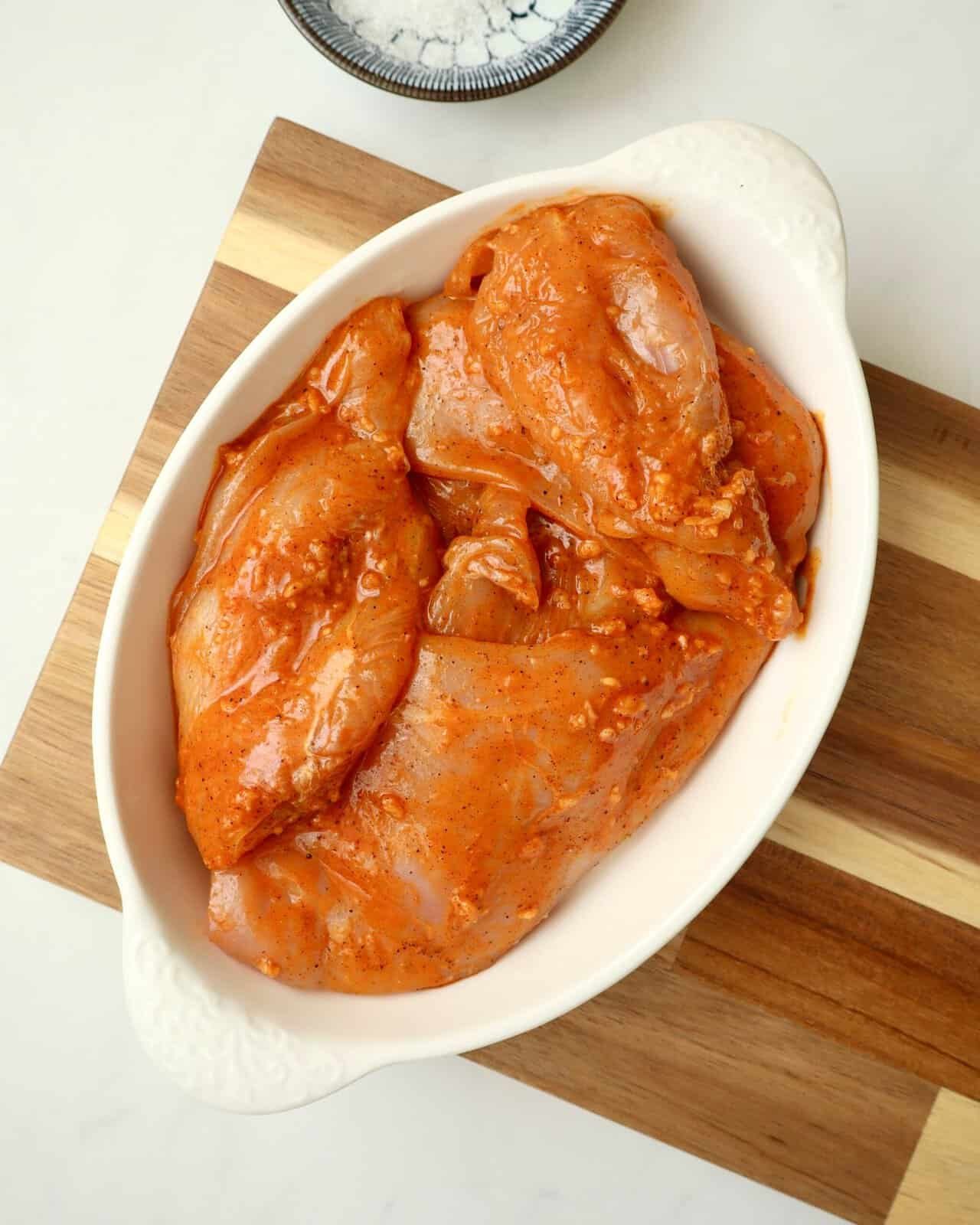 Raw chicken breasts marinating in a white bowl with herbs and seasoning