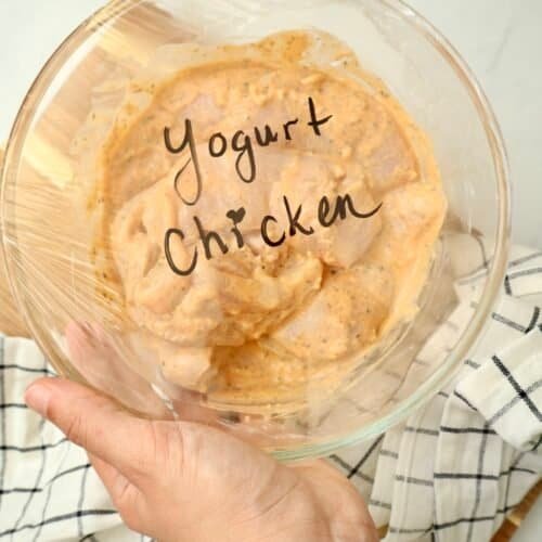Raw chicken coated in garlic yogurt marinade in a clear glass bowl, covered with plastic wrap labeled “yogurt chicken,” with a hand holding the side of the bowl