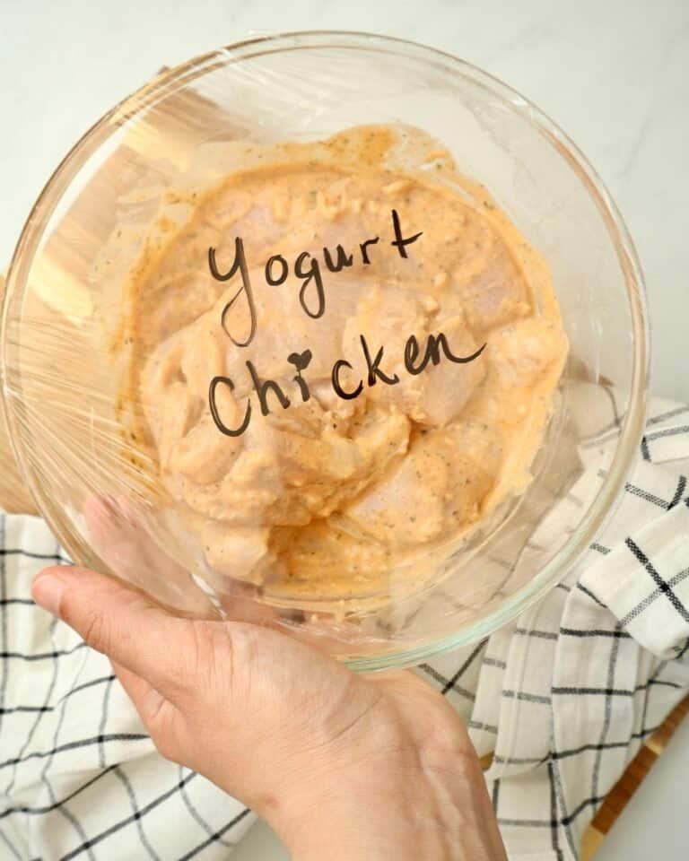 Raw chicken coated in garlic yogurt marinade in a clear glass bowl, covered with plastic wrap labeled “yogurt chicken,” with a hand holding the side of the bowl