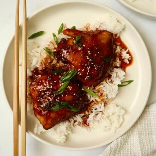 Grilled chicken thighs with spicy Asian sauce served over steamed white rice on a white plate.