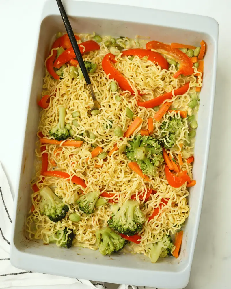 Instant ramen noodle stir-fry with broccoli, red bell peppers, and carrots in a white baking dish.